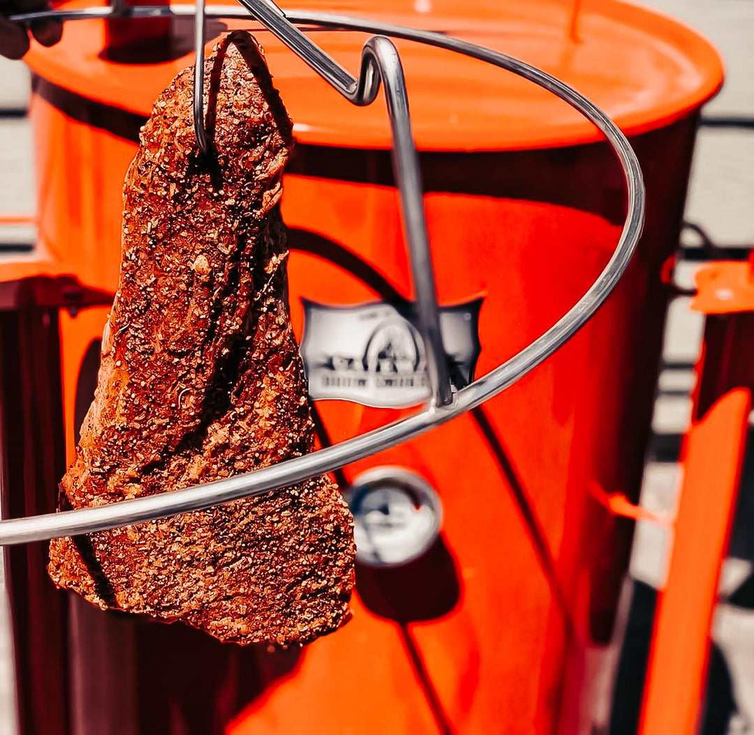 A cut of beef on the Rib Hanger Kit with a 55 gallon Gateway Drum Smoker in the background